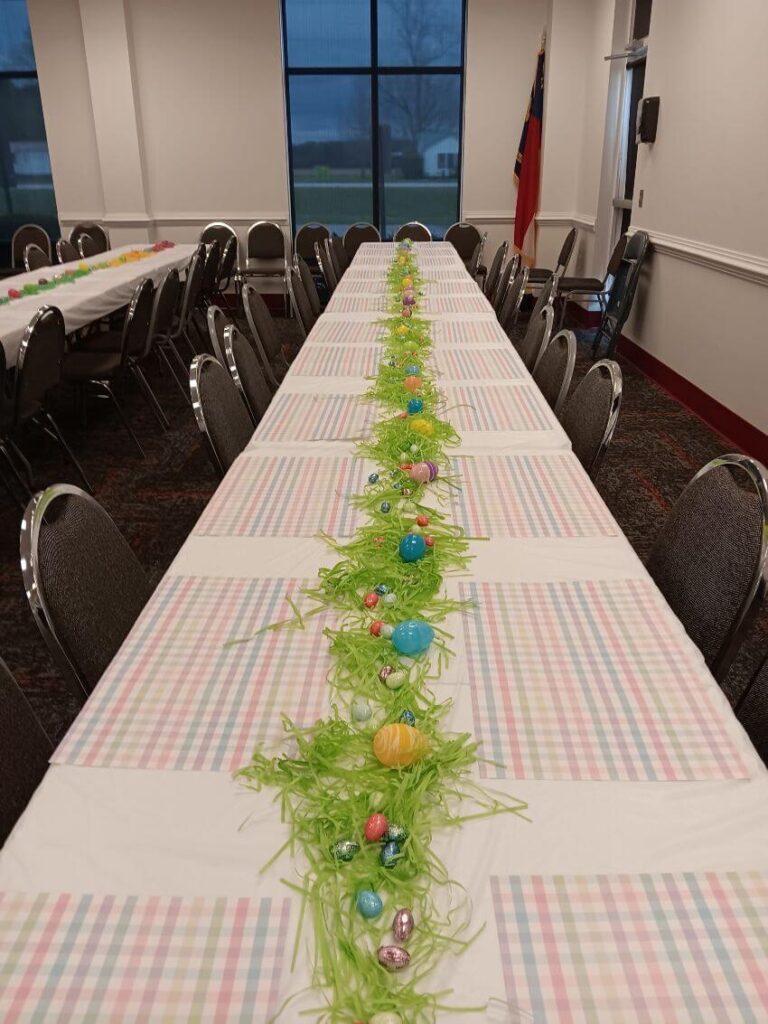 Long row of white tables with grass and easter eggs centerpiece